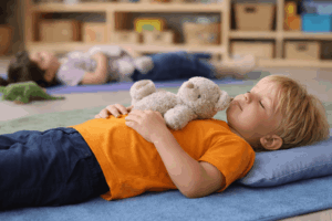 Kind im Kindergarten liegt entspannt mit Kuscheltier auf dem Bauch und macht eine Atemübung zur Entspannung im Kita-Alltag